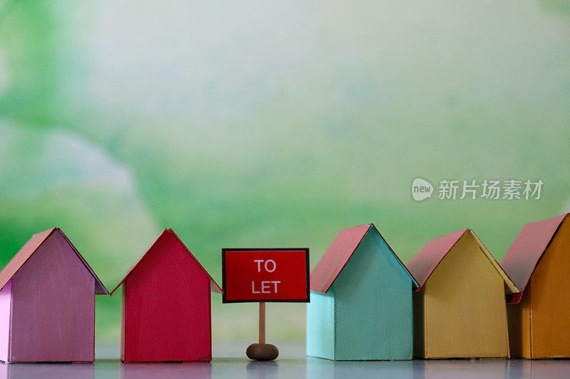 特写图像的'To Let'标志在中间排多色纸板房子，彩色的海滩小屋，重点在前景，斑杂的绿色背景，家庭财务，房地产和假日储蓄概念